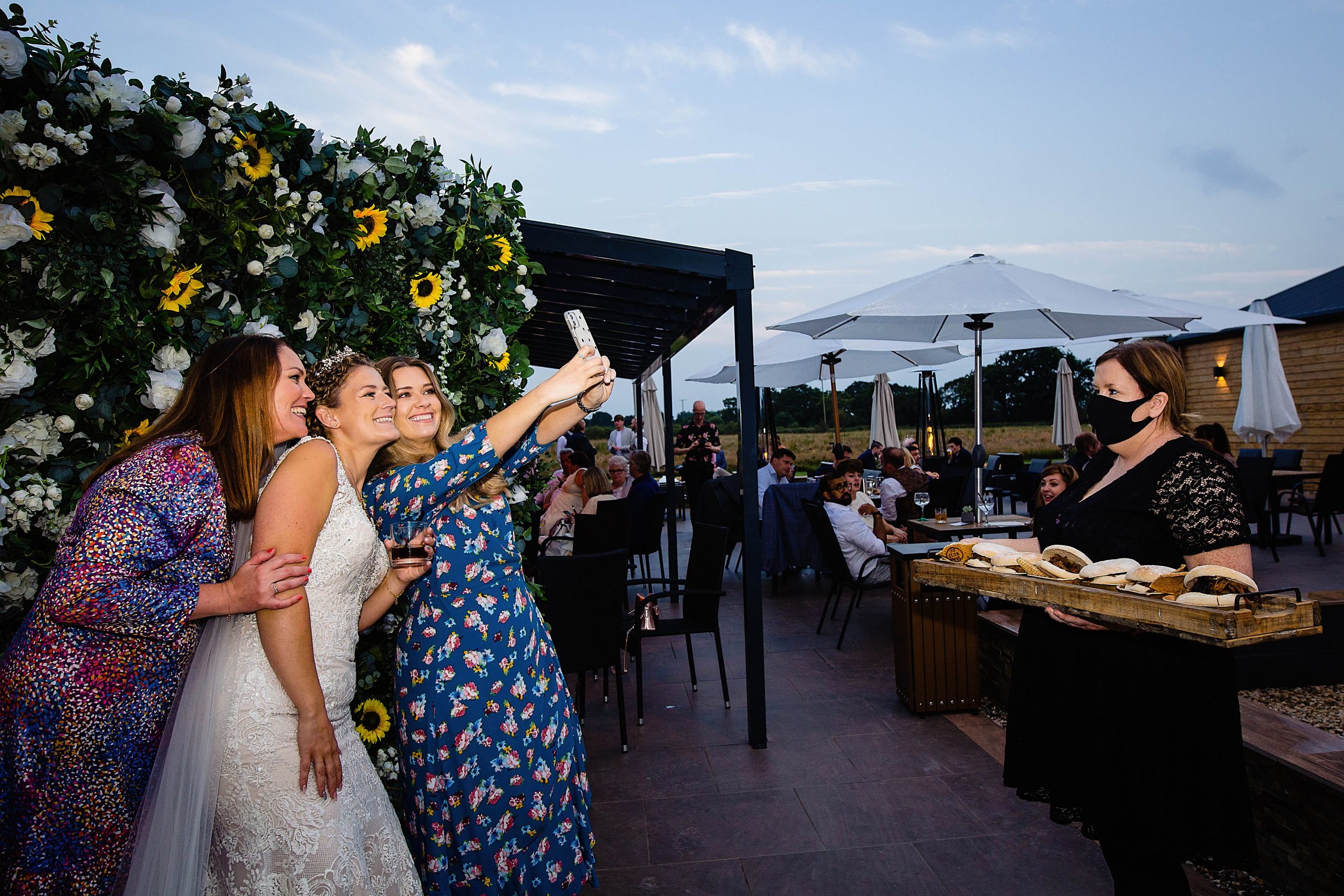 bride and wedding guests take a selfie in front of a flower wall whilst a waitress waits to pass out burgers. woodstock weddings and events wedding photography by emma and rich